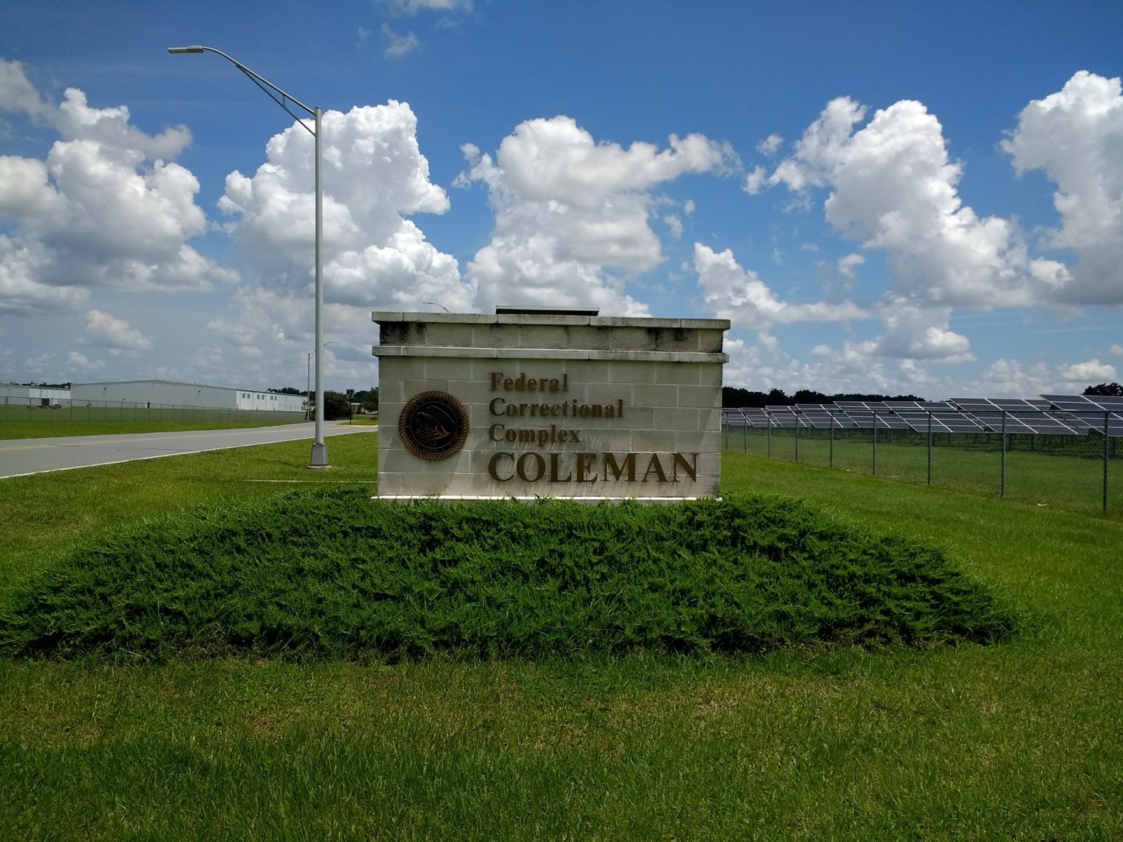A stone entrance sign reading Federal Correctional Complex Coleman on a grassy lawn beside a road, with solar panels behind a fence