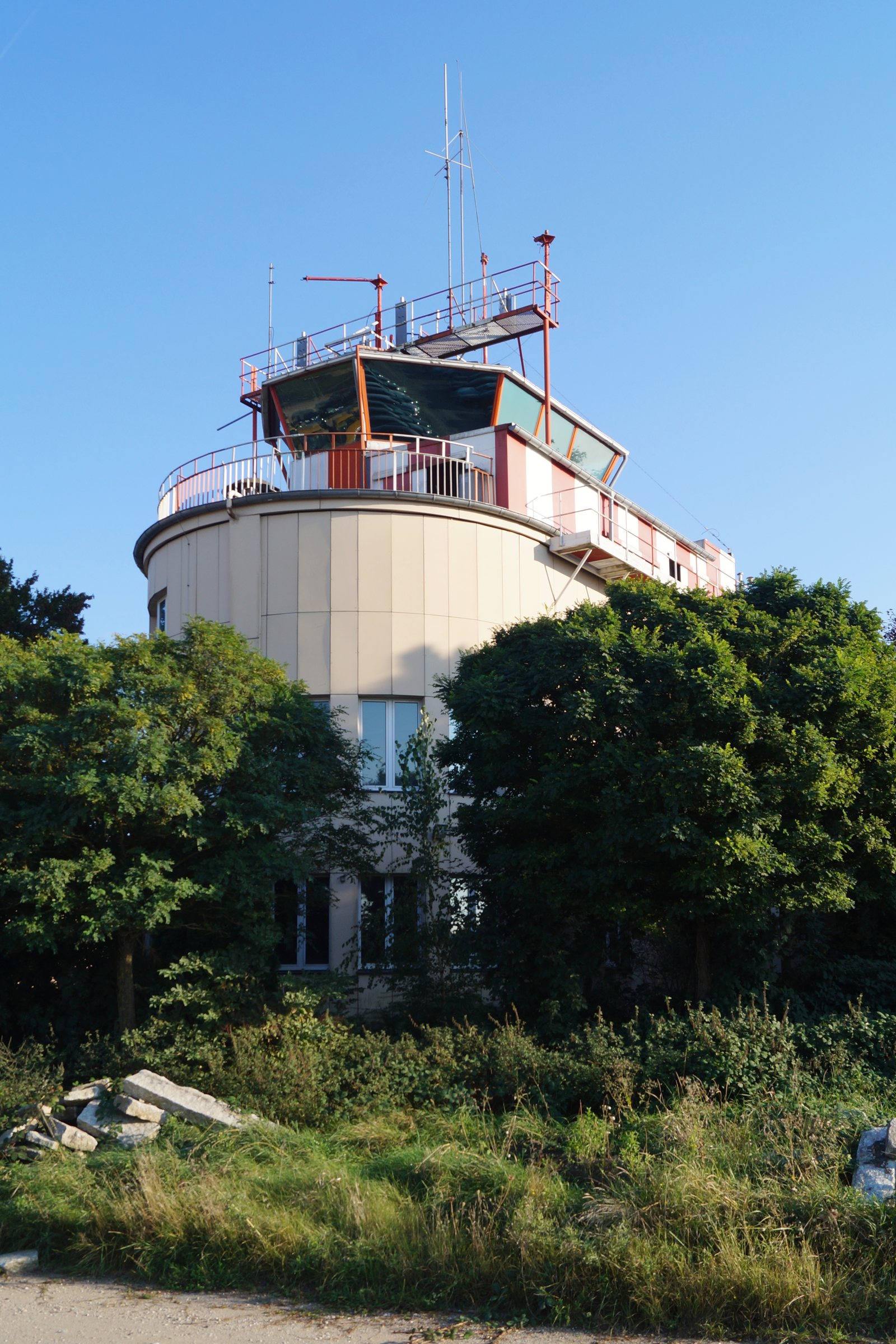 An empty airfield control tower with a rounded base and a glass cab, partly hidden by trees and overgrown grass