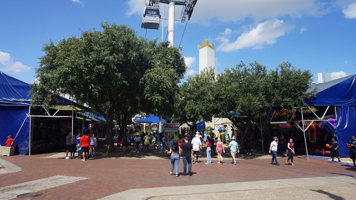 Fairgoers walking past game booths under blue tents on the State Fair of Texas Midway